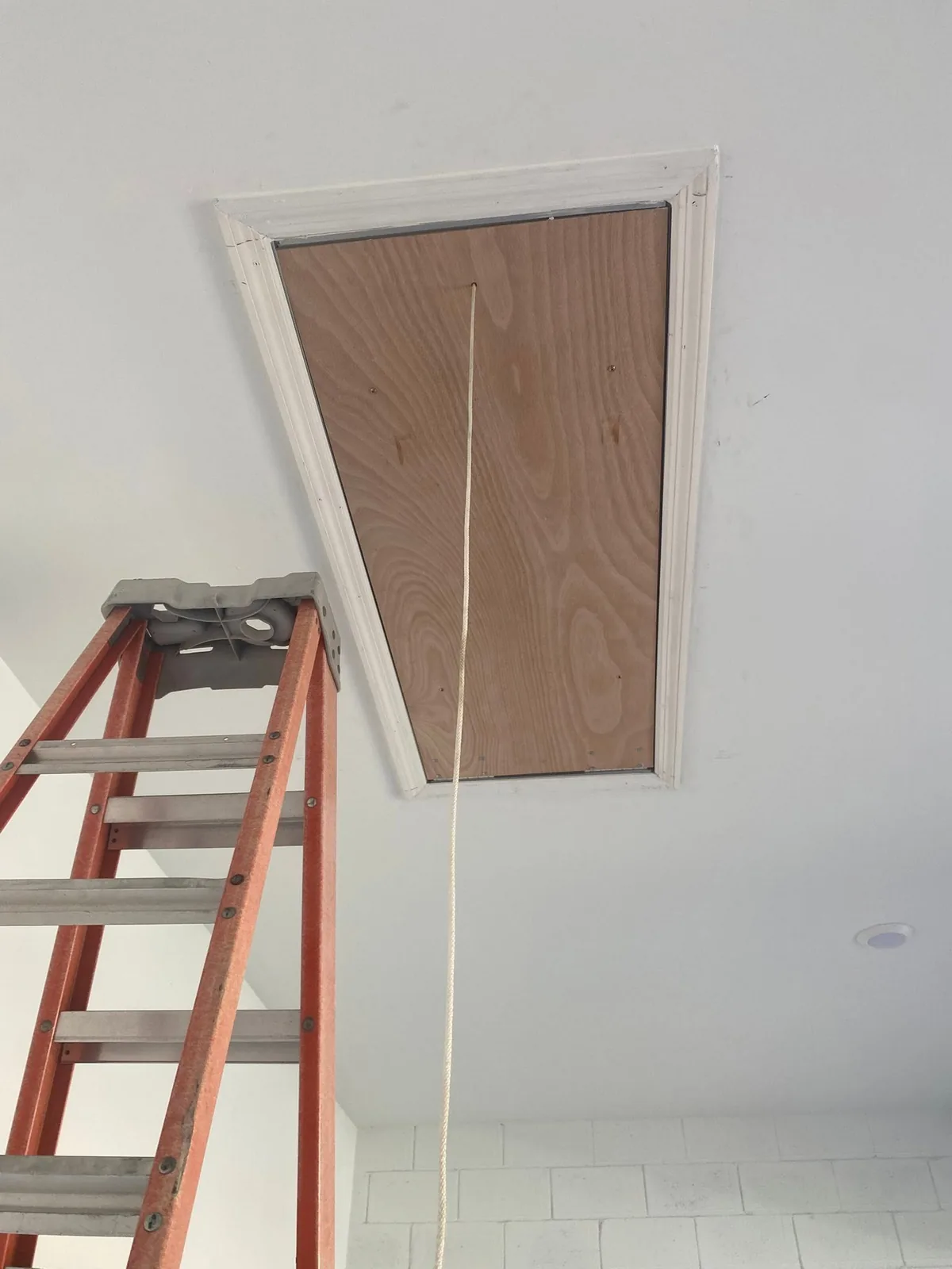 Wood ceiling access panel with white trim and a pull cord, with a ladder below.
